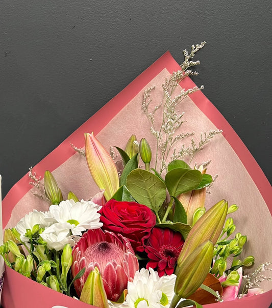 Elegant bouquet of red rose, pink protea, white daisies, lilies, and greenery wrapped in pink paper