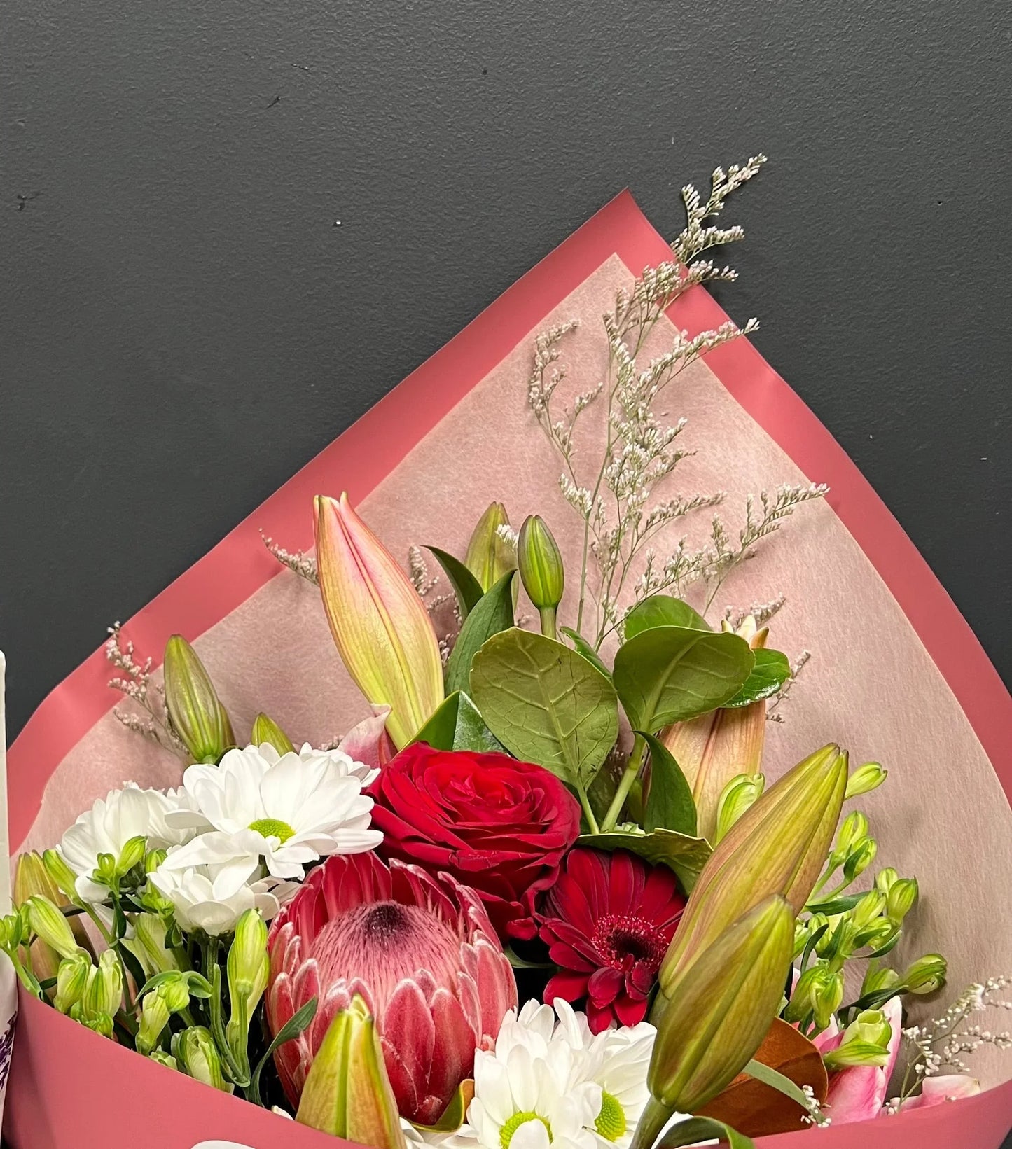 Elegant bouquet of red rose, pink protea, white daisies, lilies, and greenery wrapped in pink paper