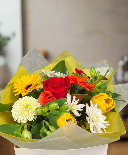 Bright mixed flower bouquet with red rose, yellow tulips, white chrysanthemums, and orange blossoms in yellow wrapping