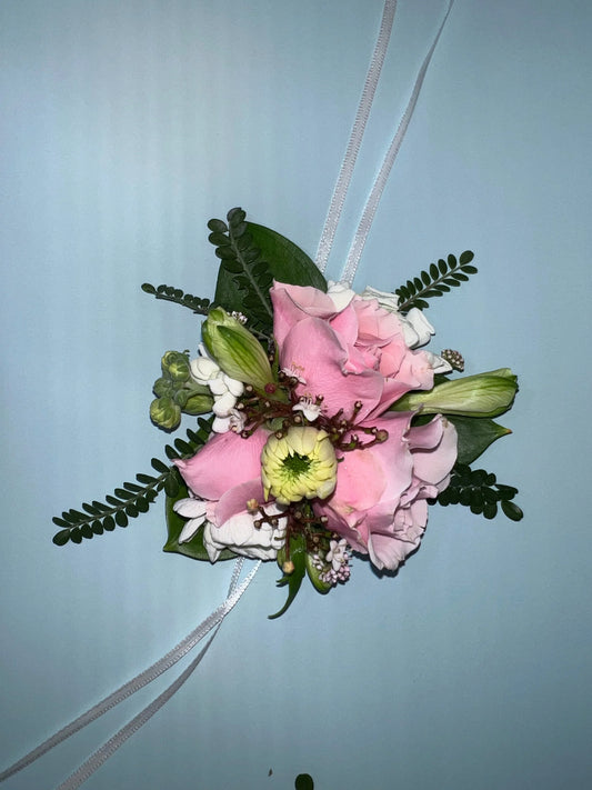 Delicate pink rose wrist corsage with white ribbon, green leaves, and small white filler flowers on light blue background