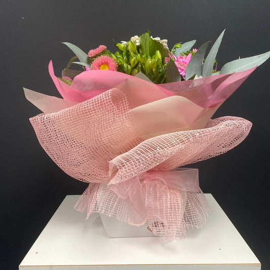 Pink and white floral bouquet with green leaves, wrapped in pink mesh and tissue paper on white pedestal