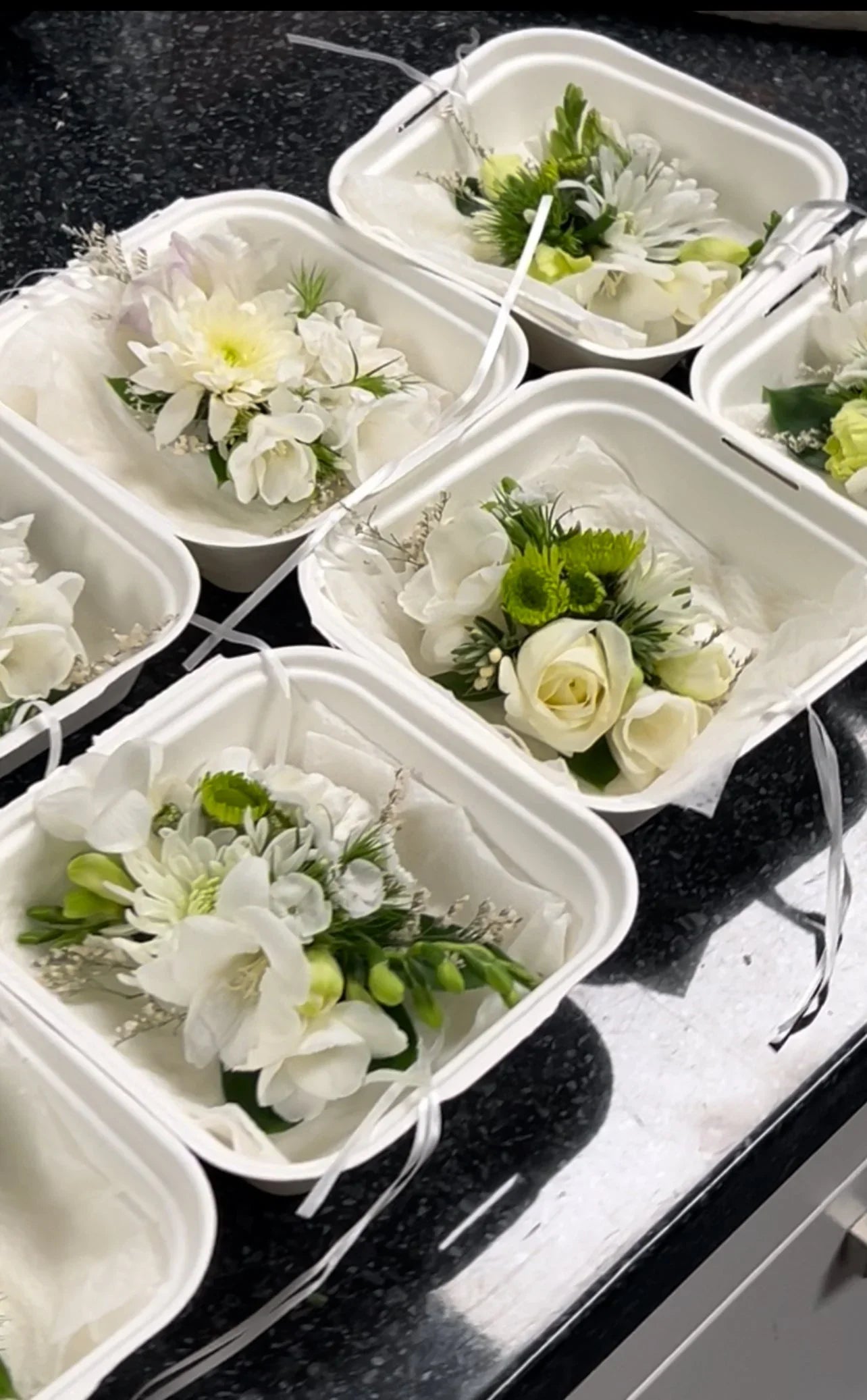White and green floral boutonnières arranged in eco-friendly white boxes on a black countertop