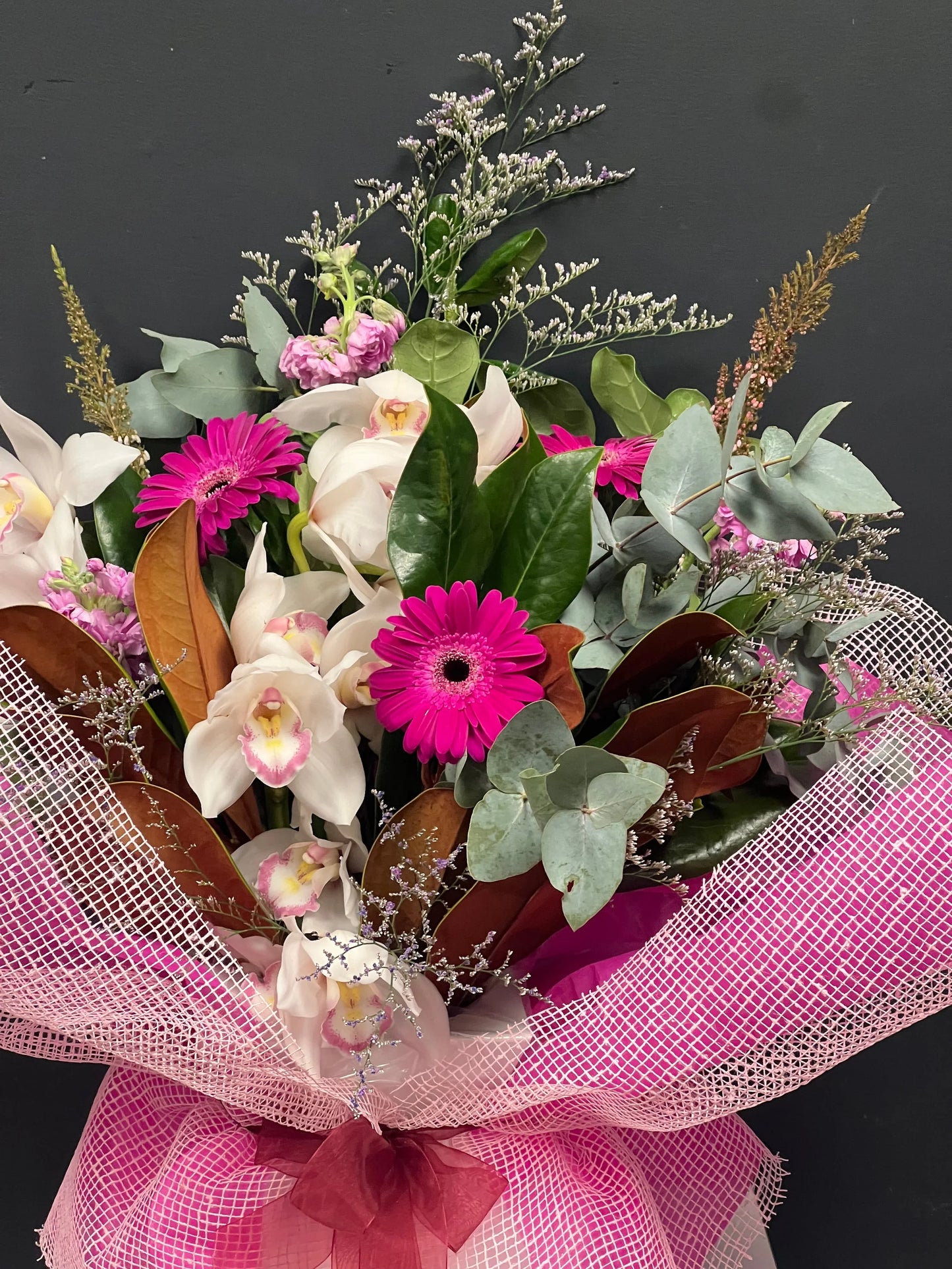 Bouquet of white orchids, pink gerbera daisies, green and brown foliage wrapped in pink mesh against black background