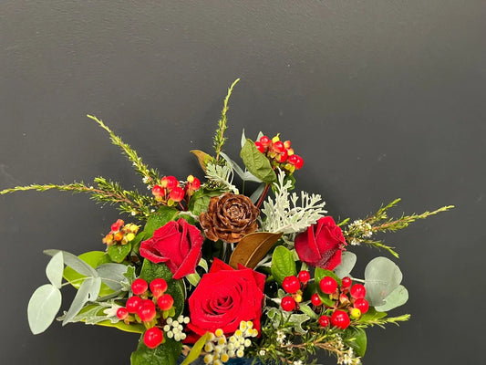Festive floral arrangement with red roses, red berries, greenery, and pinecone against a dark background