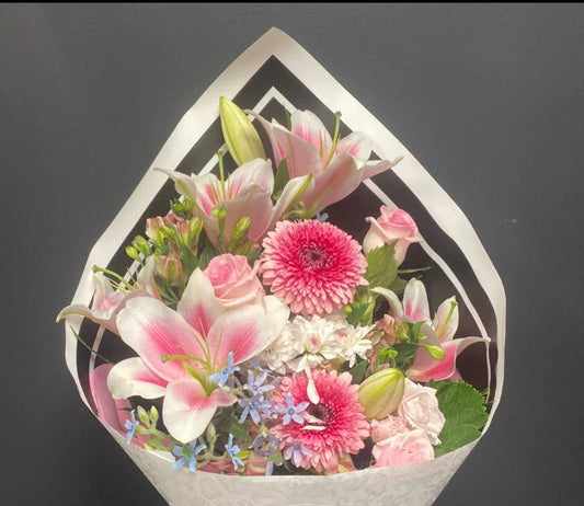 Elegant bouquet with pink lilies, gerberas, roses, and blue accent flowers wrapped in white paper against dark background