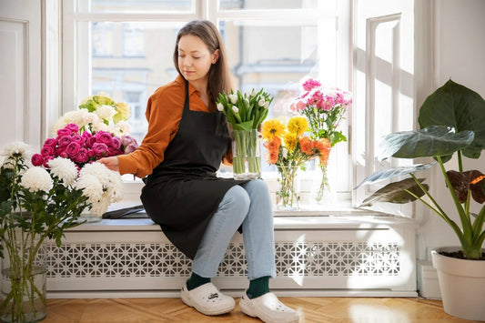 Florist woman in black apron arranging white and pink flowers near a sunlit window with green potted plant