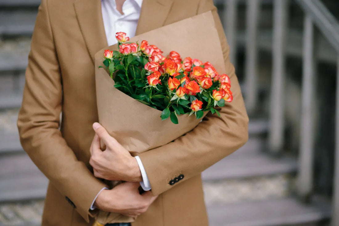 Person in brown jacket holding bouquet of orange and yellow roses wrapped in brown paper