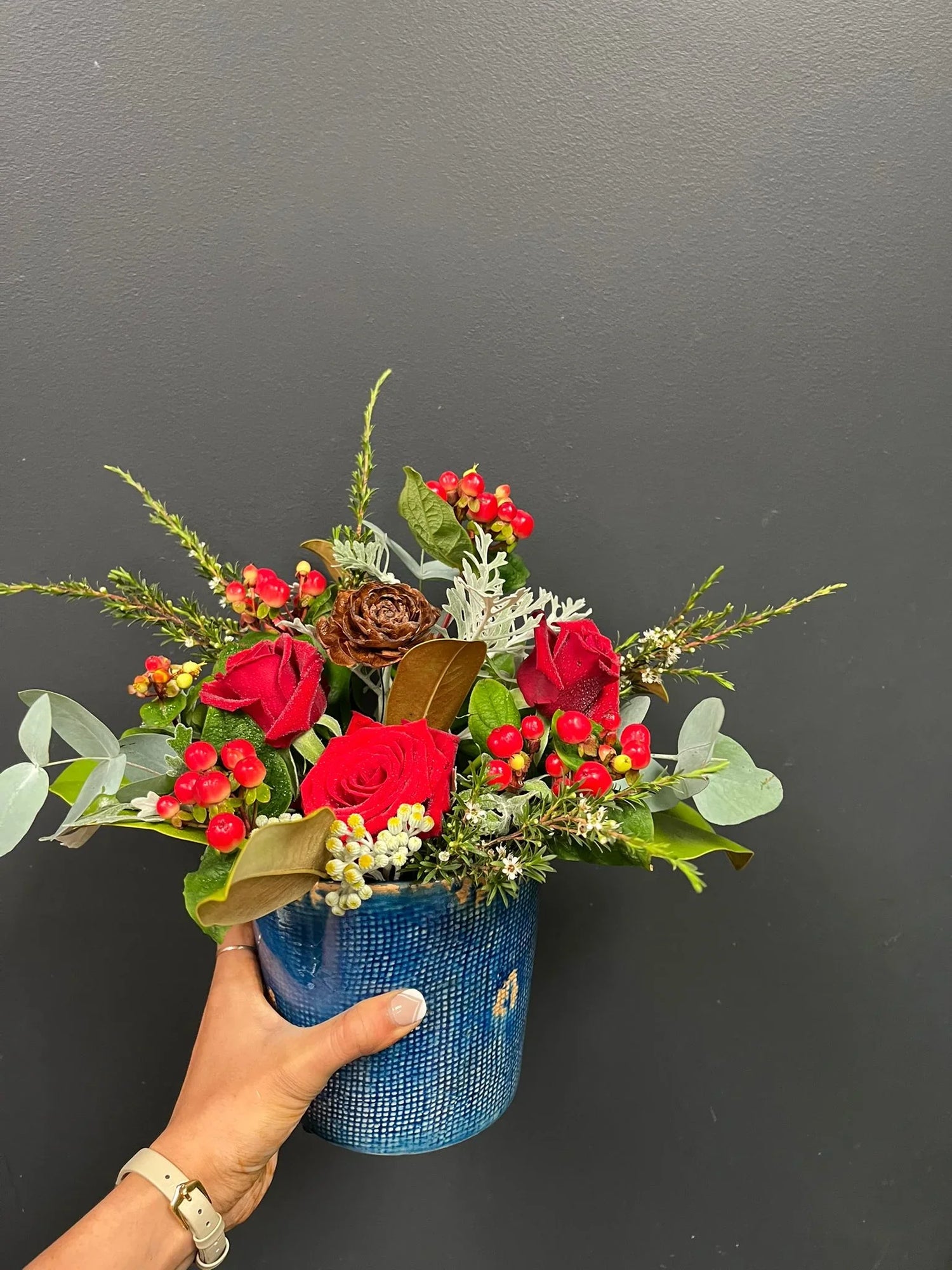 Hand holding blue vase with red roses, red berries, greenery, and pinecone against gray wall