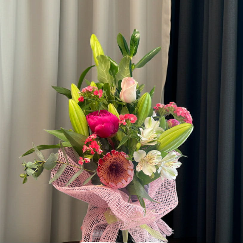 Flower bouquet with pink rose, protea, lilies, peonies, and greenery wrapped in pink mesh against a dark background